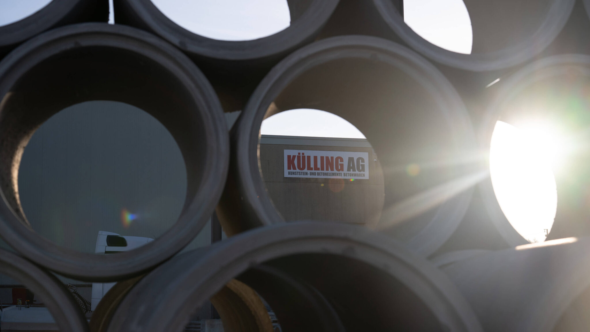 Stapel großer Betonrohre vor einem Gebäude mit dem Schild „KÜLLING AG“ im Gegenlicht