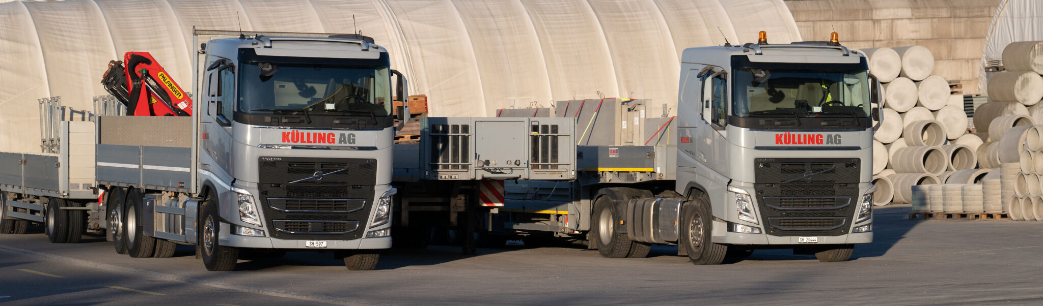 Zwei silberne KÜLLING-AG-Lkw mit Anhängern auf einem Industriegelände, neben gestapelten Betonrohren