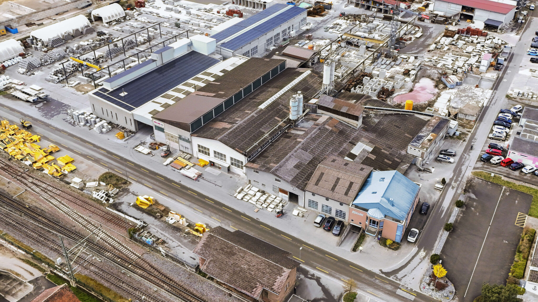 Luftaufnahme eines Industrie- und Lagergeländes mit Hallen, Materialstapeln und Bahngeleisen