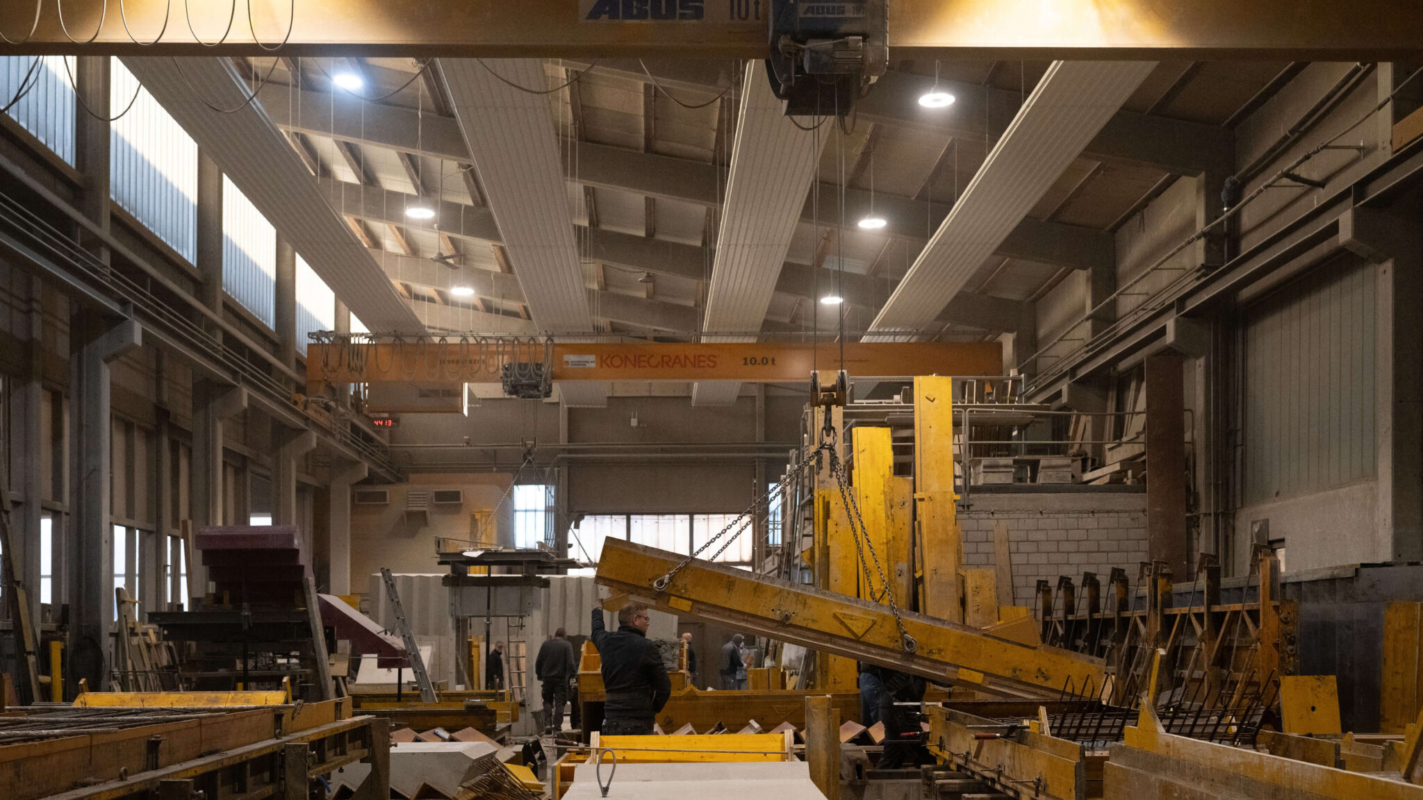 Industriehalle mit Kranen und Arbeitern beim Hantieren mit großen Metallteilen