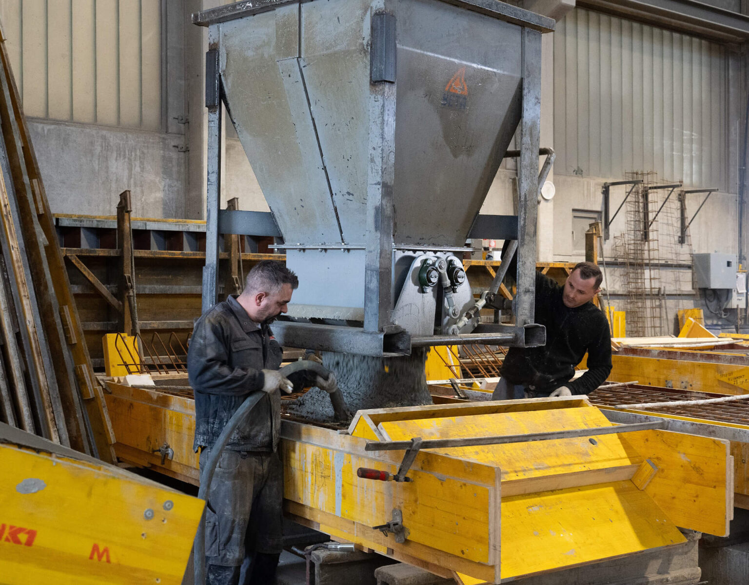 Zwei Arbeiter füllen Beton in eine gelbe Schalung in einer Werkhalle